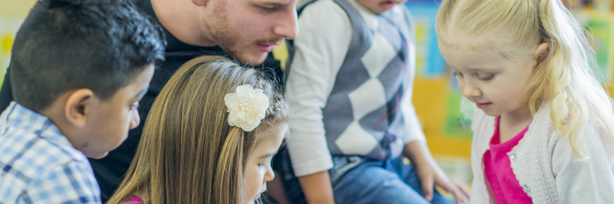 An educator is sitting on the floor with 4 preschoolers, looking at the book