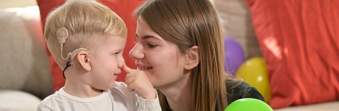 A female adult is sitting beside a toddler who is wearing hearing implants. An adult is looking at the child's eyes and smiling.