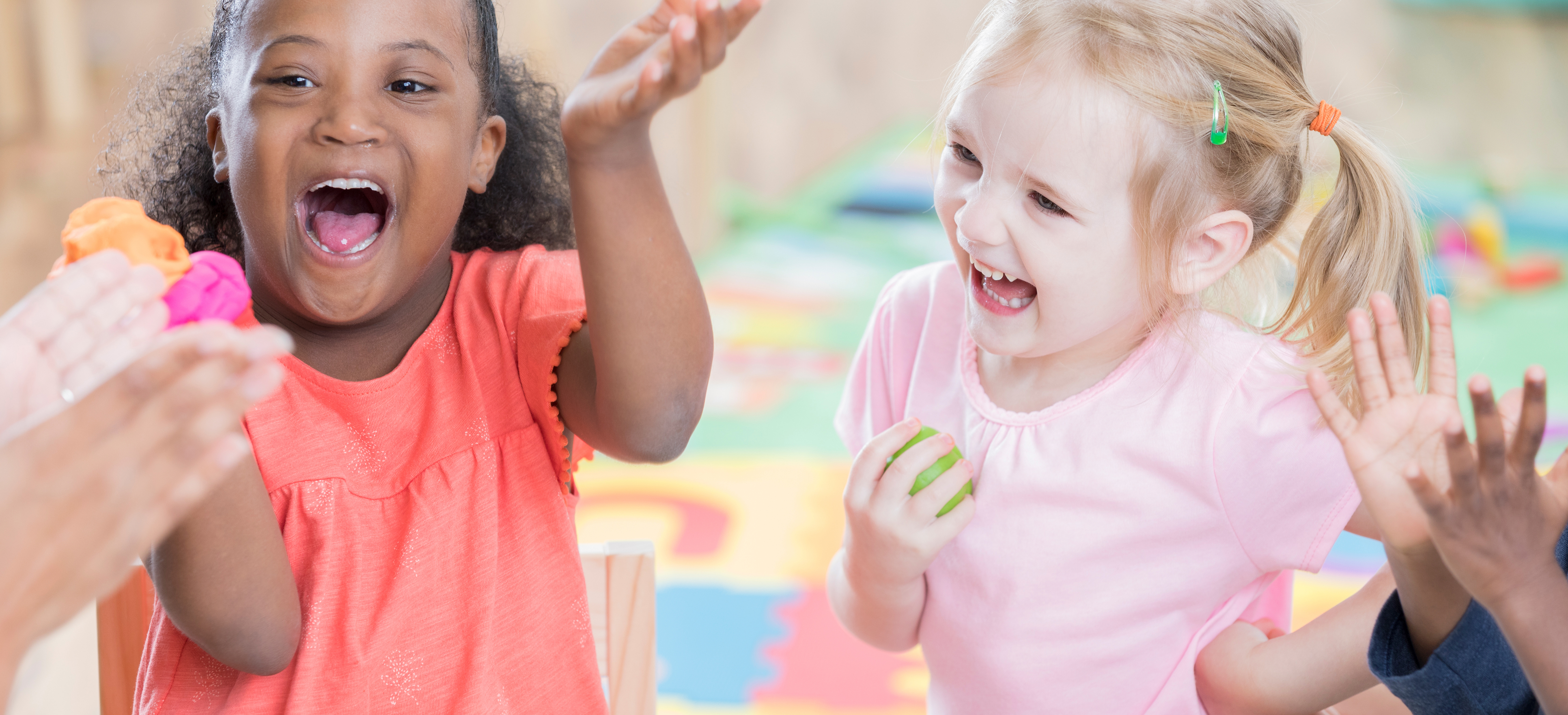 Two preschoolers are looking up at the educators clapping hands. Both children are laughing. The one is holding two balls of playdough while the other one is holding one.