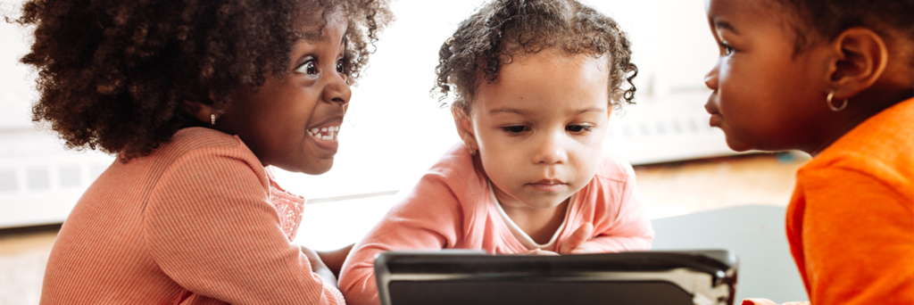 three preschool girls are sitting at a table; one is looking through an I-Pad, two others are looking at each other 